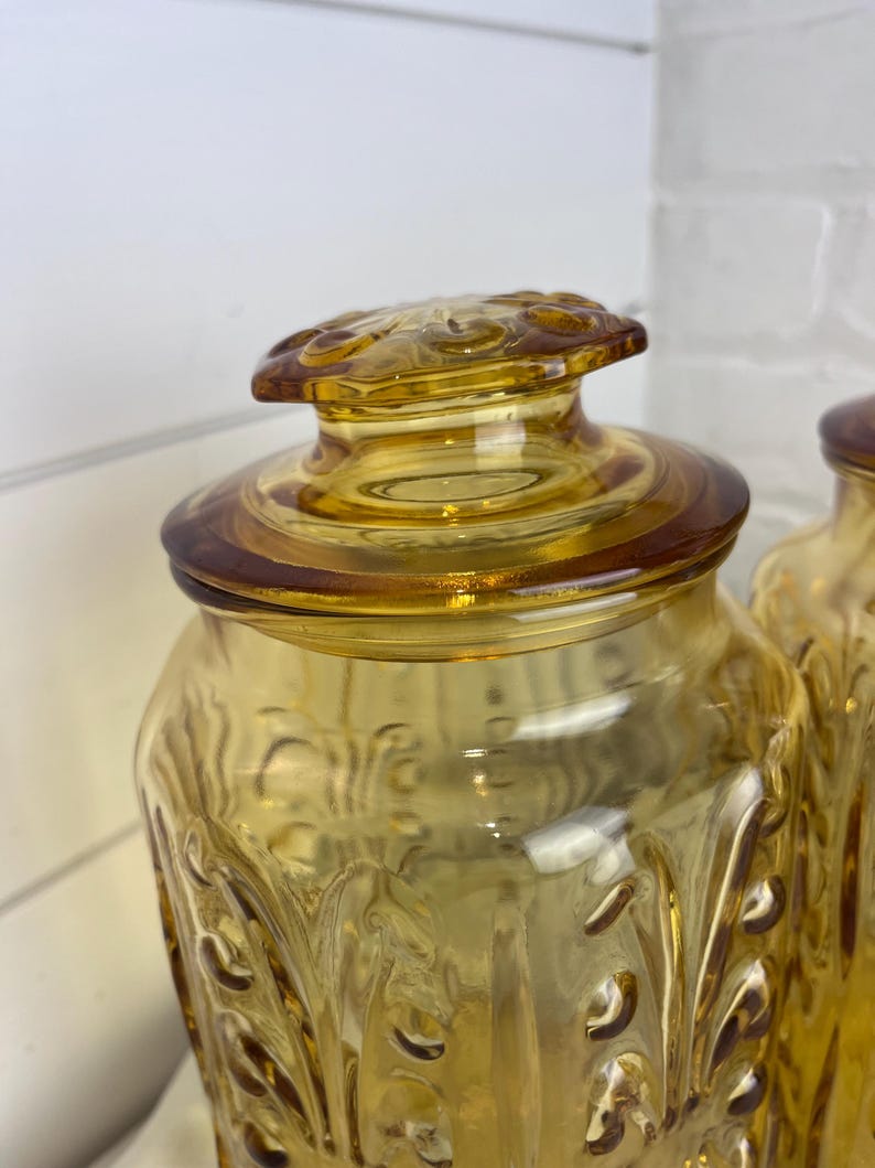 May include: Amber-colored glass jar with a decorative lid. The jar has a textured, embossed pattern and a matching lid with a floral design. The jar is likely used for storage and adds a vintage touch to any kitchen.