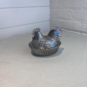 May include: Iridescent glass hen-shaped dish with a detailed rooster lid. The dish has a ruffled edge and a textured surface, reflecting a spectrum of colors. A decorative kitchenware item.