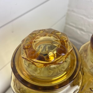 May include: Close-up of two amber-colored glass jars with decorative lids. The jars have a textured surface and a floral pattern. The lids are hexagonal with a decorative rim. The jars are likely for storage or display.