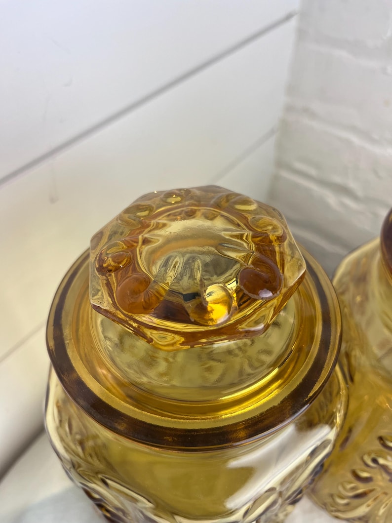 May include: Close-up of two amber-colored glass jars with decorative lids. The jars have a textured surface and a floral pattern. The lids are hexagonal with a decorative rim. The jars are likely for storage or display.