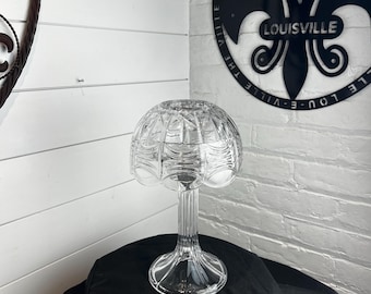 May include: A clear glass candle holder featuring a dome-shaped top with a faceted, geometric pattern and a tall, slender, vertically ribbed stem that flares out at the base. The glass is transparent and reflects light across its textured surface.