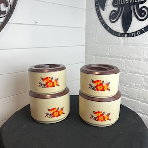 May include: Two sets of stacked cylindrical food containers with cream-colored bodies and dark brown lids. Each container features a floral pattern with bright orange and red flowers and green leaves printed on the front side of the exterior.