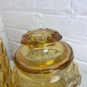 May include: Amber-colored glass jar with a decorative lid. The jar has a square shape with embossed floral patterns. The lid is also amber-colored and features a floral design. The jar is likely vintage and suitable for kitchen storage.