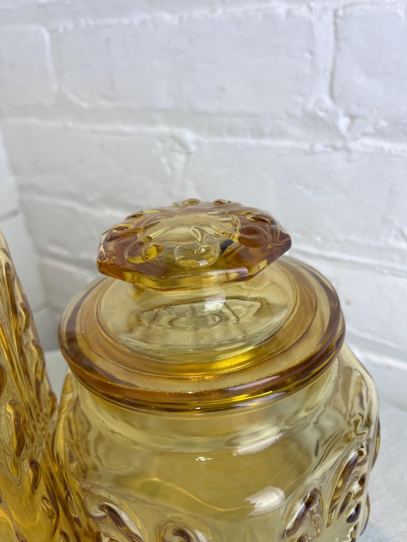 May include: Amber-colored glass jar with a decorative lid. The jar has a square shape with embossed floral patterns. The lid is also amber-colored and features a floral design. The jar is likely vintage and suitable for kitchen storage.