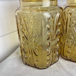 May include: Two amber-colored glass jars with lids. The jars have a decorative, embossed floral pattern and a square shape. The lids are round and fit snugly on top. The jars are likely for kitchen storage.