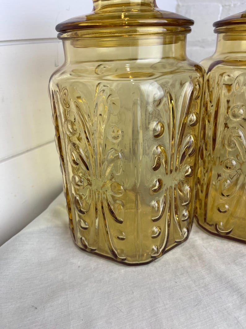 May include: Two amber-colored glass jars with lids. The jars have a decorative, embossed floral pattern and a square shape. The lids are round and fit snugly on top. The jars are likely for kitchen storage.