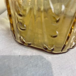 May include: A vintage amber-colored glass container with a textured, patterned design. The glass has a glossy finish and is slightly translucent. The container's base is rounded, and the sides feature vertical ridges and decorative elements.