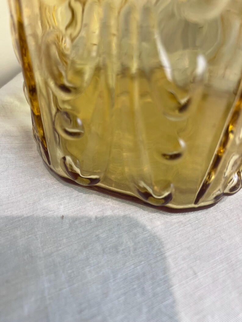 May include: A vintage amber-colored glass container with a textured, patterned design. The glass has a glossy finish and is slightly translucent. The container's base is rounded, and the sides feature vertical ridges and decorative elements.