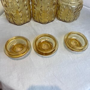 May include: A collection of amber-colored glass jars and lids. The jars have decorative embossed patterns and varying shapes, including tall, cylindrical, and hexagonal designs. The lids are round and fit snugly on the jars.