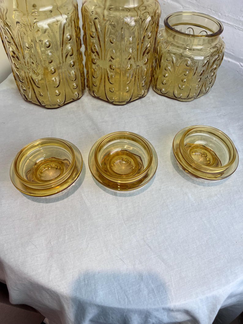 May include: A collection of amber-colored glass jars and lids. The jars have decorative embossed patterns and varying shapes, including tall, cylindrical, and hexagonal designs. The lids are round and fit snugly on the jars.