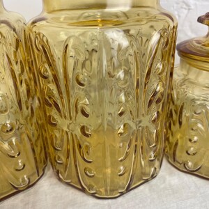 May include: Amber-colored glass jars with lids. The jars have a decorative, embossed pattern and a rounded shape. The lids are also amber-colored and fit snugly on top. The jars are likely for storage or display.