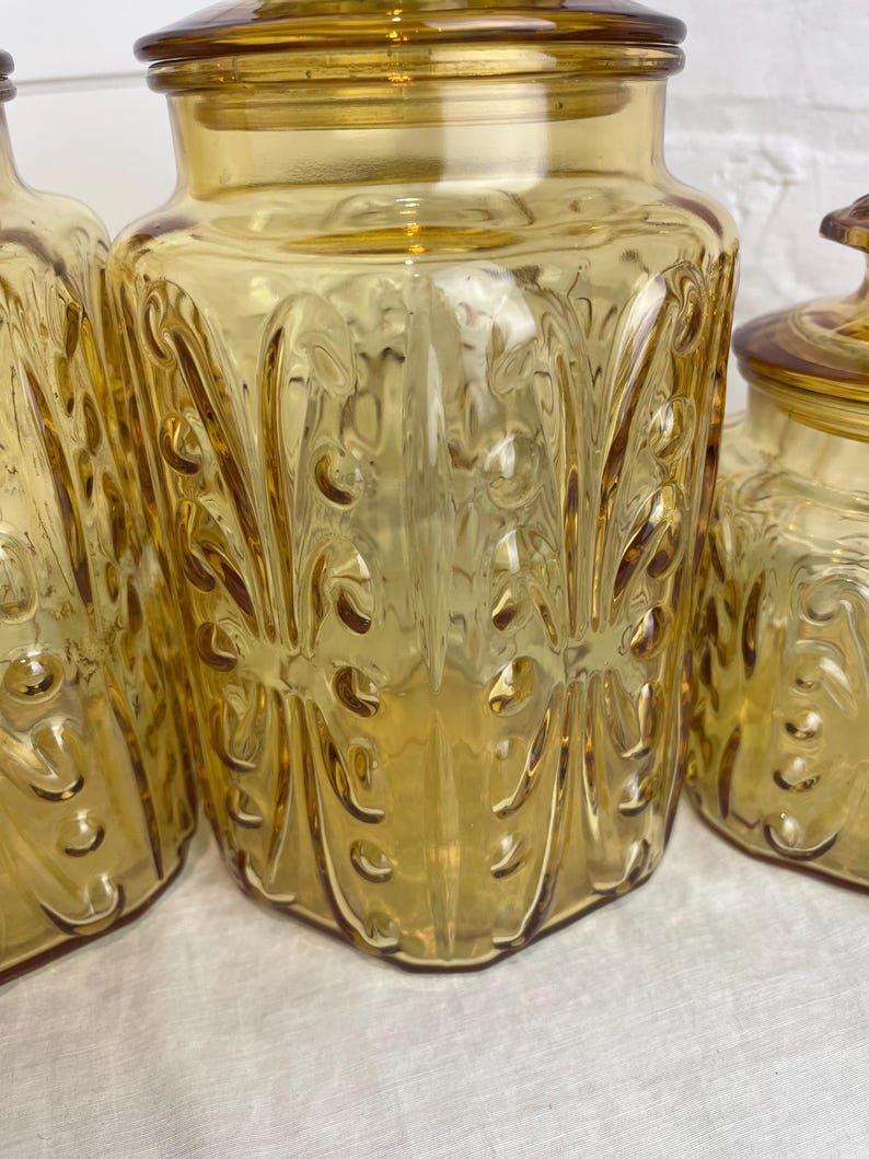 May include: Amber-colored glass jars with lids. The jars have a decorative, embossed pattern and a rounded shape. The lids are also amber-colored and fit snugly on top. The jars are likely for storage or display.