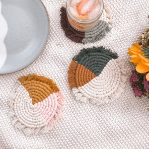 May include: Three handmade macrame coasters in a variety of colors, including pink, brown, and green. The coasters are round and have a fringe border. They are sitting on a white crocheted surface.