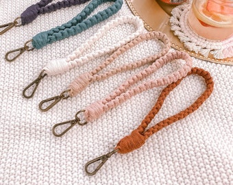 May include: A collection of macrame wristlets in various colors, including burgundy, black, gray, teal, cream, and brown. Each wristlet features a braided design and a bronze-colored clasp. The wristlets are displayed on a white knitted surface.