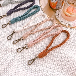 May include: A collection of macrame wristlets in various colors, including burgundy, black, gray, teal, cream, and brown. Each wristlet features a braided design and a bronze-colored clasp. The wristlets are displayed on a white knitted surface.