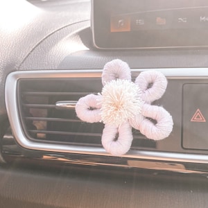 May include: A lavender and white flower-shaped car air freshener hanging from a car's air vent. The air freshener is made of fabric and has a pom-pom in the center.