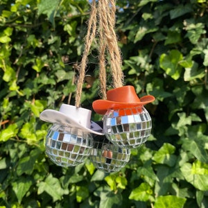 May include: Three silver disco ball ornaments with a red and white cowboy hat on each. The ornaments are hanging from a twine string.