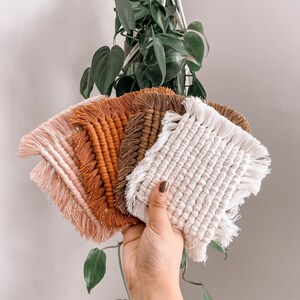 May include: Five handmade macrame coasters in various shades of brown, orange, and pink with fringe edges. The coasters are held in a hand against a white wall.