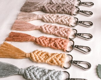 May include: A set of eight colorful macrame keychains with silver key rings. The keychains are made with a variety of colors, including pink, orange, yellow, blue, and gray. Each keychain has a tassel at the end.