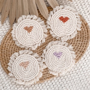 May include: Four macrame coasters with a cream-colored base and fringed edges. Each coaster features a different heart design in shades of peach, rust, and lavender. The coasters are arranged on a woven tray with a dried palm leaf in the background.