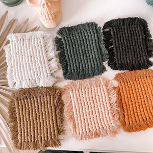 May include: Six handmade macrame coasters in various colors, including white, black, brown, pink, and orange. Each coaster has a square shape with fringed edges.