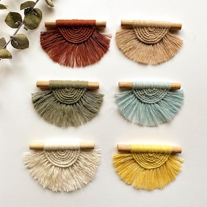 Könnte beinhalten: Sechs kleine Wandbehänge aus Makramee mit Holzstäben. Die Behänge sind in verschiedenen Farben, darunter Rost, Beige, Grün, Hellblau, Creme und Gelb.
