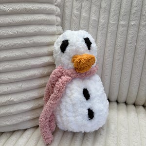 Könnte beinhalten: Ein handgemachter, plüschiger Schneemann mit weißem Körper, schwarzen Augen und Kohleknöpfen. Er hat eine orangefarbene Nase und einen rosa Schal. Der Schneemann ist aus weichem, strukturiertem Garn gefertigt und eine gemütliche Winterdekoration.