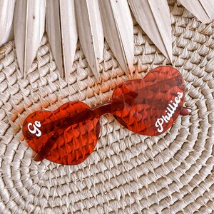 May include: Red heart-shaped sunglasses with white text that says "Go Phillies" on a woven straw background.
