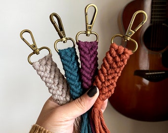 May include: Four colorful macrame keychains with tassels. The keychains are made with a braided rope and have a gold metal clasp. The keychains are in the colors beige, teal, burgundy, and orange.