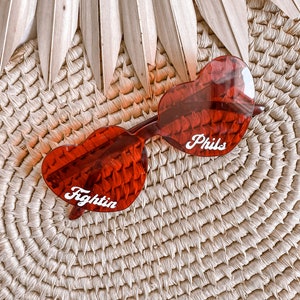 May include: Red heart-shaped sunglasses with white text that says "Fightin" on one lens and "Phils" on the other lens. The sunglasses are on a woven straw surface.