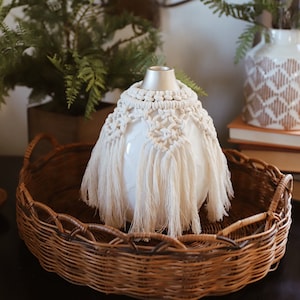 May include: A white ceramic diffuser with a macrame cover and tassels sits on a brown wicker tray. The diffuser is surrounded by green plants.