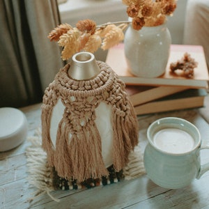 May include: A white ceramic diffuser with a brown macrame cover and tassels sits on a wooden table with a light blue mug and a stack of books. Dried flowers in a white vase are in the background.