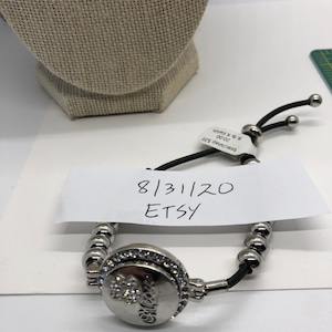 May include: A silver bracelet with a round, jeweled charm that says "Believe". The bracelet has a black cord and silver beads. A white tag with the date 8/31/20 and the word "Etsy" is visible.