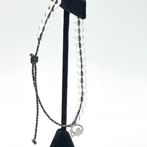 May include: A clear plastic bracelet with a silver charm in the shape of a fish. The bracelet is on a black display stand.