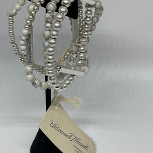 May include: A set of five bracelets with white and silver beads. The bracelets are stacked on a black stand with a tag that reads "Universal Thread Goods Co."