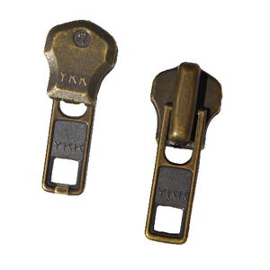 Könnte beinhalten: Zwei antike goldene Reißverschluss-Zipper mit einem schwarzen Kunststoff-Schieber und dem Text "YKK" auf jedem.
