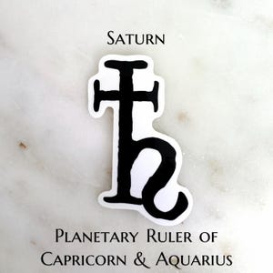 May include: A black and white sticker featuring the astrological symbol for Saturn. The word "SATURN" is printed above the symbol, and the text "PLANETARY RULER OF CAPRICORN & AQUARIUS" is printed below.