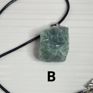 May include: A green stone pendant on a black cord necklace. The stone is rough and has a natural, irregular shape. The pendant is labeled with the letter "B".
