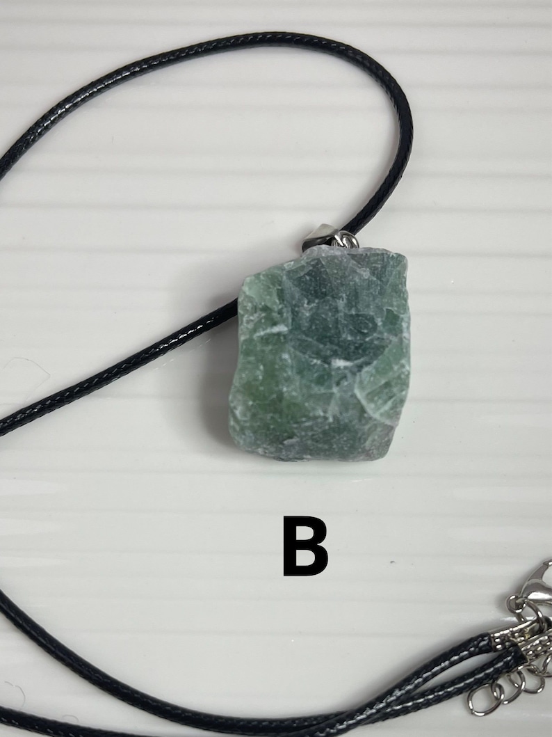 May include: A green stone pendant on a black cord necklace. The stone is rough and has a natural, irregular shape. The pendant is labeled with the letter "B".