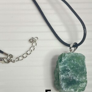 May include: A green crystal pendant necklace with a black cord and silver chain. The pendant is a rough, natural-looking crystal with a green and white color.