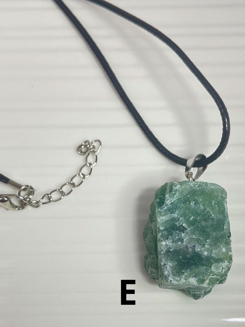 May include: A green crystal pendant necklace with a black cord and silver chain. The pendant is a rough, natural-looking crystal with a green and white color.