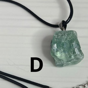 May include: A green crystal pendant on a black cord necklace with a silver clasp. The pendant is a rough, natural-looking stone with a light green color.