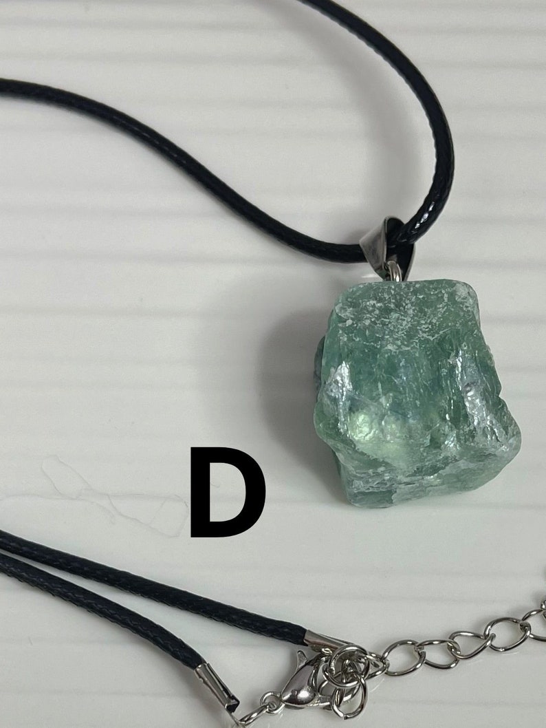 May include: A green crystal pendant on a black cord necklace with a silver clasp. The pendant is a rough, natural-looking stone with a light green color.