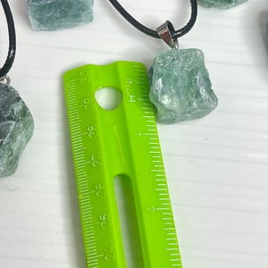 May include: A close-up of a green gemstone pendant necklace with a black cord. The pendant is about 1 inch in size and is next to a ruler.