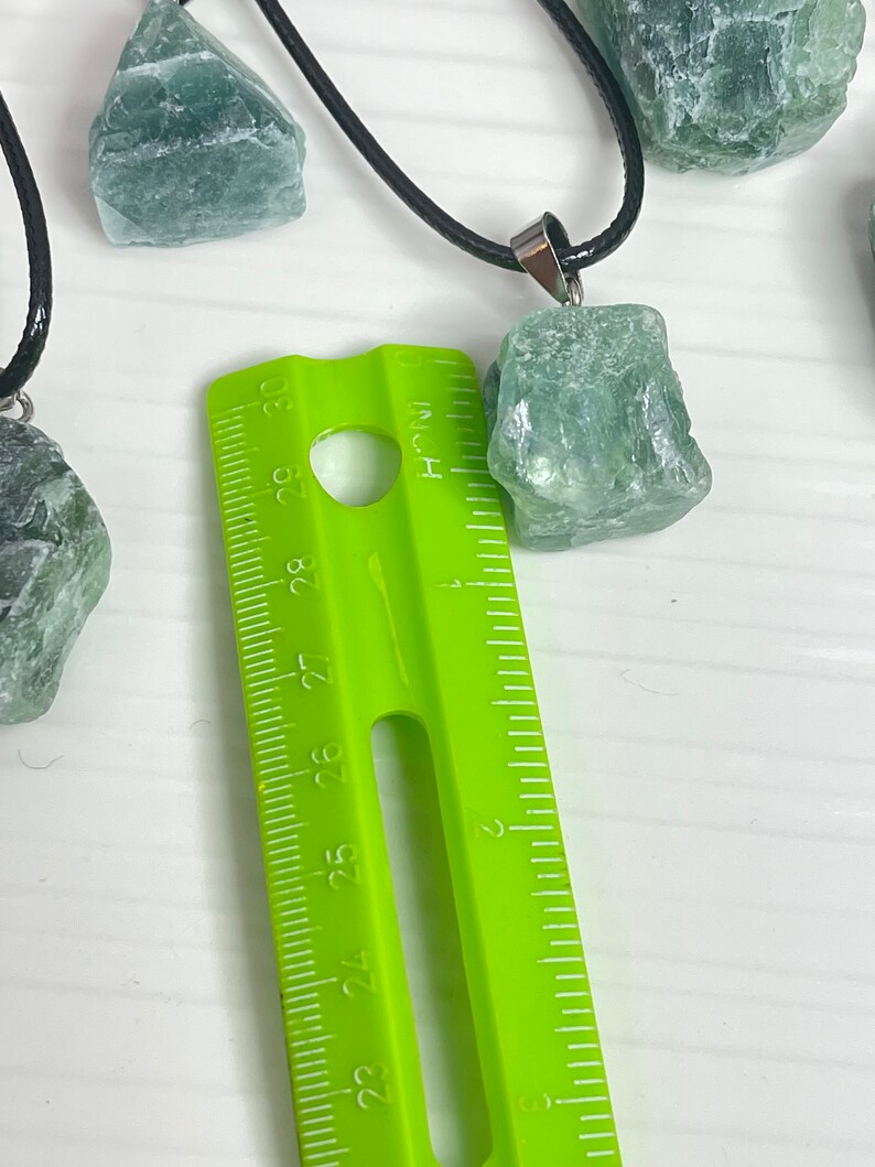 May include: A close-up of a green gemstone pendant necklace with a black cord. The pendant is about 1 inch in size and is next to a ruler.