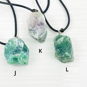 May include: Three necklaces with different green and purple crystal pendants. The pendants are all rough-cut and have a natural, organic look. The necklaces are labeled J, K, and L.