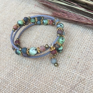 May include: A double-wrap bracelet with green, brown, and red beads, accented with gold beads and a gold charm. The bracelet is on a burlap background.