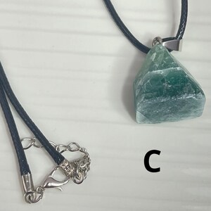 May include: A green gemstone pendant on a black cord necklace. The pendant is a rough, irregular shape and has a silver clasp. The necklace has a silver lobster clasp.