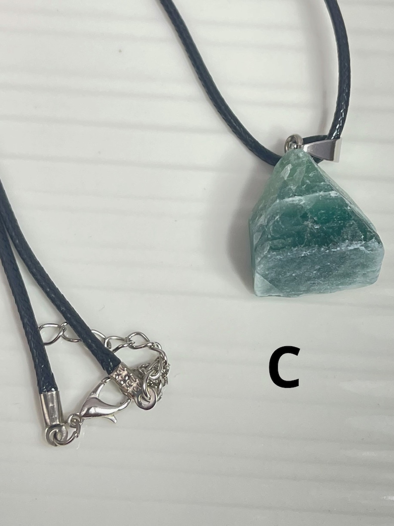 May include: A green gemstone pendant on a black cord necklace. The pendant is a rough, irregular shape and has a silver clasp. The necklace has a silver lobster clasp.