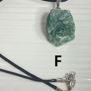 May include: A green stone pendant on a black cord necklace. The pendant is a rough, irregular shape with a silver bail. The necklace has a silver clasp. The letter "F" is printed on a white surface below the necklace.